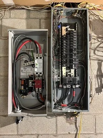 Open automatic transfer switch beside an open breaker panel on a block wall