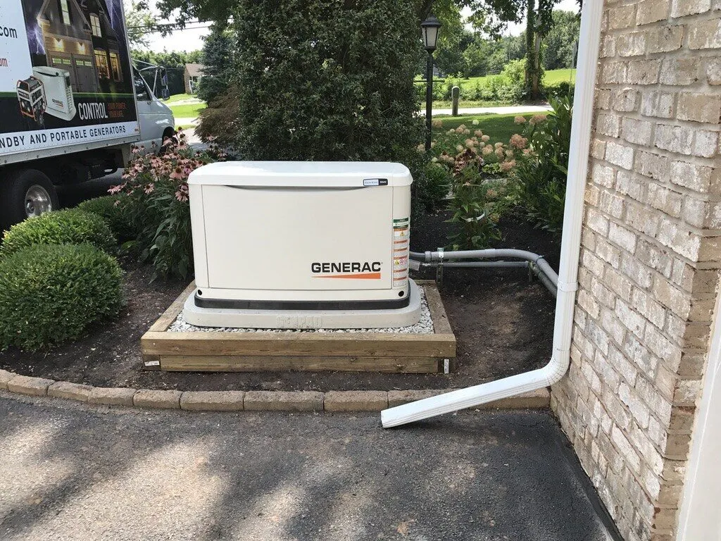 Generac standby generator on a timber-edged pad tucked between shrubs and a brick wall