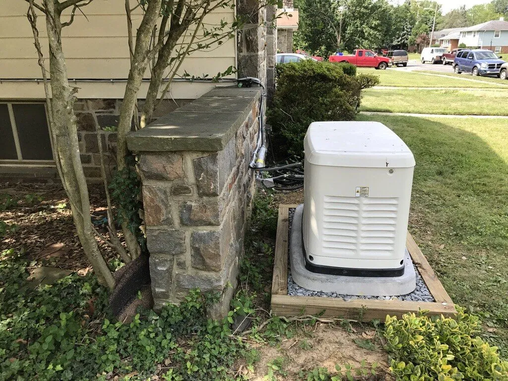 Generac standby generator on a framed gravel pad next to a stone pillar and shrubs