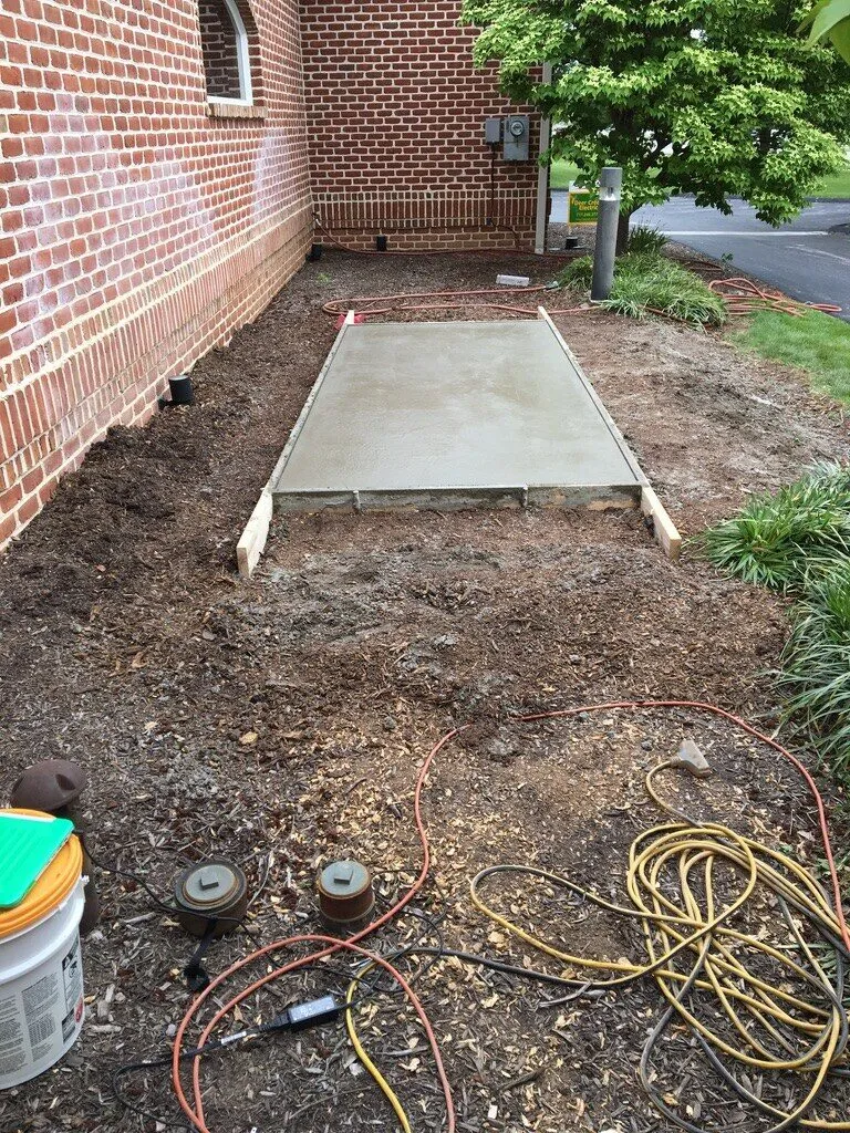 Freshly poured concrete generator pad along a brick wall, with tools and cords around it